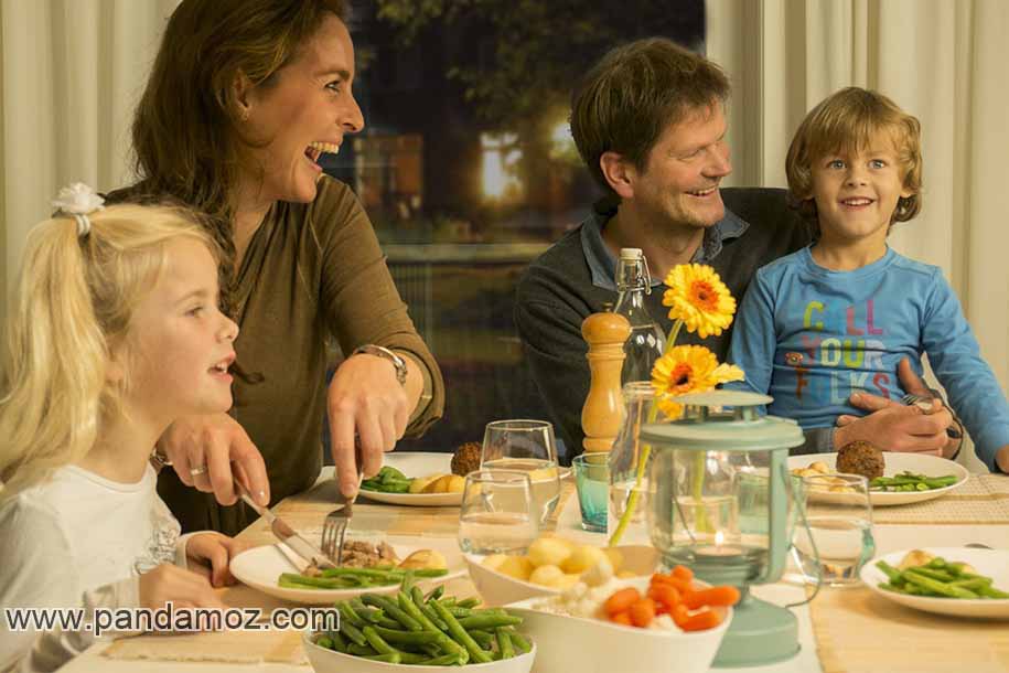 عکس خانواده شاد شامل زن و شوهر،دختر و پسر. در تصویر اعضای خانواده با شادی در کنار هم پشت میز غذاخوری نشسته و خنده کنان و در حال صحبت مشغول خوردن هستند. پسر در بغل پدر و دختربچه در کنار مادر است