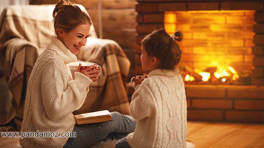 عکس یک خانه گرم و شومینه روشن دارای آتش و زن و دختر بچه ایی که فنجان قهوه در دست دارند و می نوشند و مبلمان خانه دیده می شود