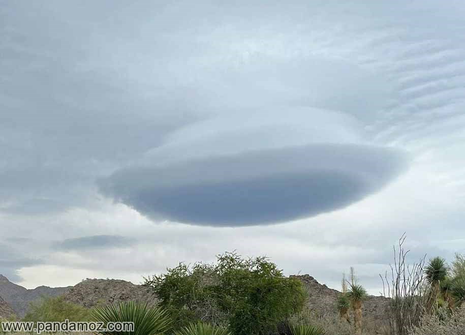 عکس یک تکه ابر در آسمان که به شکلی زیبا و به صورت گرد است و در بالای کوه و دامنه های کوه قرار گرفته است. گیاهان روی زمین در تصویر دیده می شوند